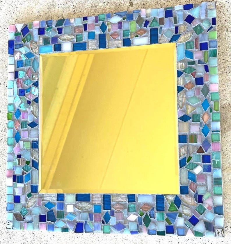 Op de afbeelding: Een vierkante spiegel met een decoratieve lijst bestaande uit veelkleurige glazen moza&iuml;ektegels in tinten blauw, groen, wit en zachtroze. Het centrale reflecterende oppervlak is glad en goudkleurig, omringd door een rand van diverse geometrische vormen.