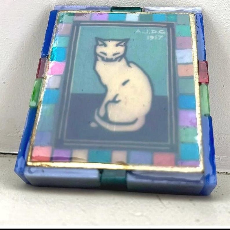 May include: A rectangular decorative tile featuring a central illustration of a seated cream-colored cat against a teal background. The image includes the text A.J.D.G 1917. The border consists of a mosaic of small, multicolored glass squares.