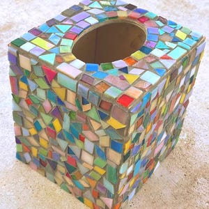 May include: A square tissue box cover decorated with a multi-colored mosaic pattern. The surface features small, irregularly shaped glass tiles in shades of red, blue, yellow, green, and purple, set into a neutral-toned grout with an oval opening.