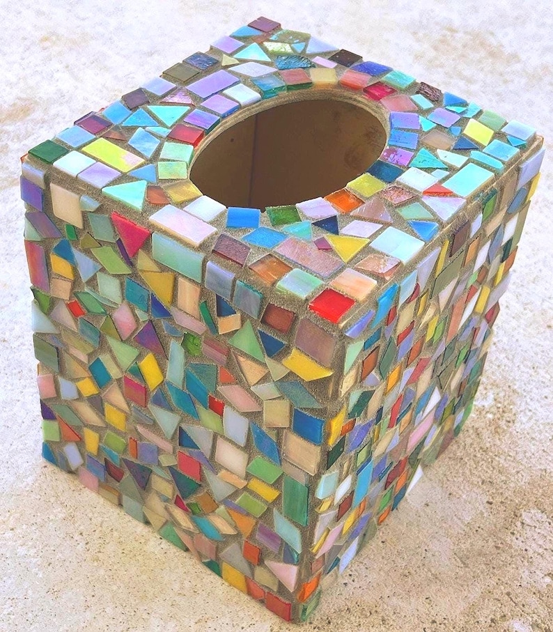May include: A square tissue box cover decorated with a multi-colored mosaic pattern. The surface features small, irregularly shaped glass tiles in shades of red, blue, yellow, green, and purple, set into a neutral-toned grout with an oval opening.