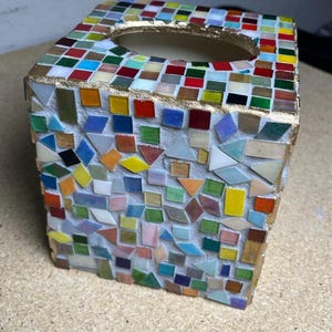 K&ouml;nnte beinhalten: Ein quadratischer Taschentuchbox-&Uuml;berzug, verziert mit einem Mosaik aus kleinen, bunten Glasfliesen in Rot, Blau, Gr&uuml;n, Gelb und Wei&szlig;. Die oberen Kanten und die ovale &Ouml;ffnung sind mit einer goldfarbenen Umrandung auf wei&szlig;em Fugenm&ouml;rtel versehen.