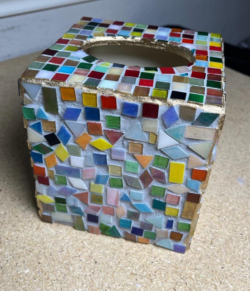 K&ouml;nnte beinhalten: Ein quadratischer Taschentuchbox-&Uuml;berzug, verziert mit einem Mosaik aus kleinen, bunten Glasfliesen in Rot, Blau, Gr&uuml;n, Gelb und Wei&szlig;. Die oberen Kanten und die ovale &Ouml;ffnung sind mit einer goldfarbenen Umrandung auf wei&szlig;em Fugenm&ouml;rtel versehen.