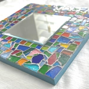 May include: A square wall mirror featuring a frame decorated with a mosaic of multi-colored glass shards in shades of blue, green, red, yellow, and pink, set in light gray grout with a light blue painted wooden edge surrounding the perimeter.