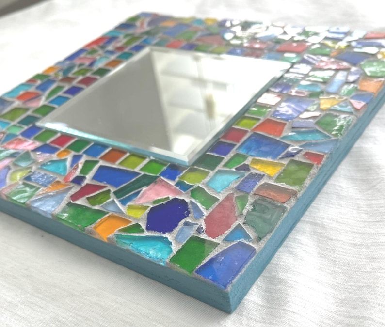 May include: A square wall mirror featuring a frame decorated with a mosaic of multi-colored glass shards in shades of blue, green, red, yellow, and pink, set in light gray grout with a light blue painted wooden edge surrounding the perimeter.