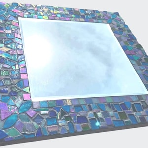 Puede incluir: Un espejo cuadrado con un marco decorativo compuesto por peque&ntilde;as piezas de vidrio en mosaico de diversas formas en tonos azules, morados y verde azulado. Las piezas est&aacute;n dispuestas en un patr&oacute;n denso con juntas visibles alrededor del espejo.