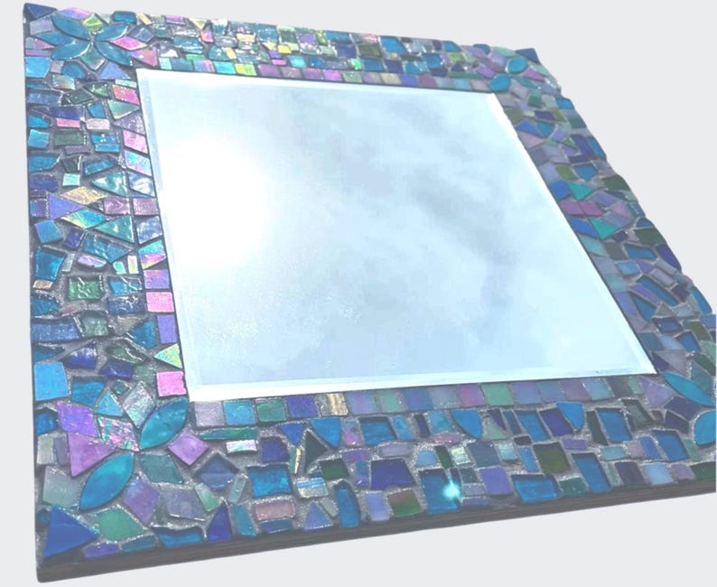 Puede incluir: Un espejo cuadrado con un marco decorativo compuesto por peque&ntilde;as piezas de vidrio en mosaico de diversas formas en tonos azules, morados y verde azulado. Las piezas est&aacute;n dispuestas en un patr&oacute;n denso con juntas visibles alrededor del espejo.