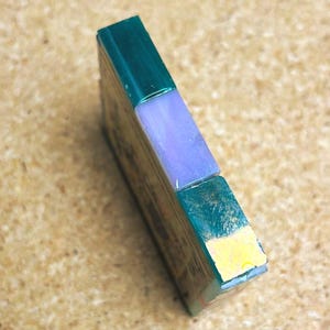 May include: A rectangular block featuring a dark green top section, a light purple middle segment, and a dark green bottom section with a small yellow accent on the lower edge. The object has a smooth, polished finish and a flat, vertical orientation.