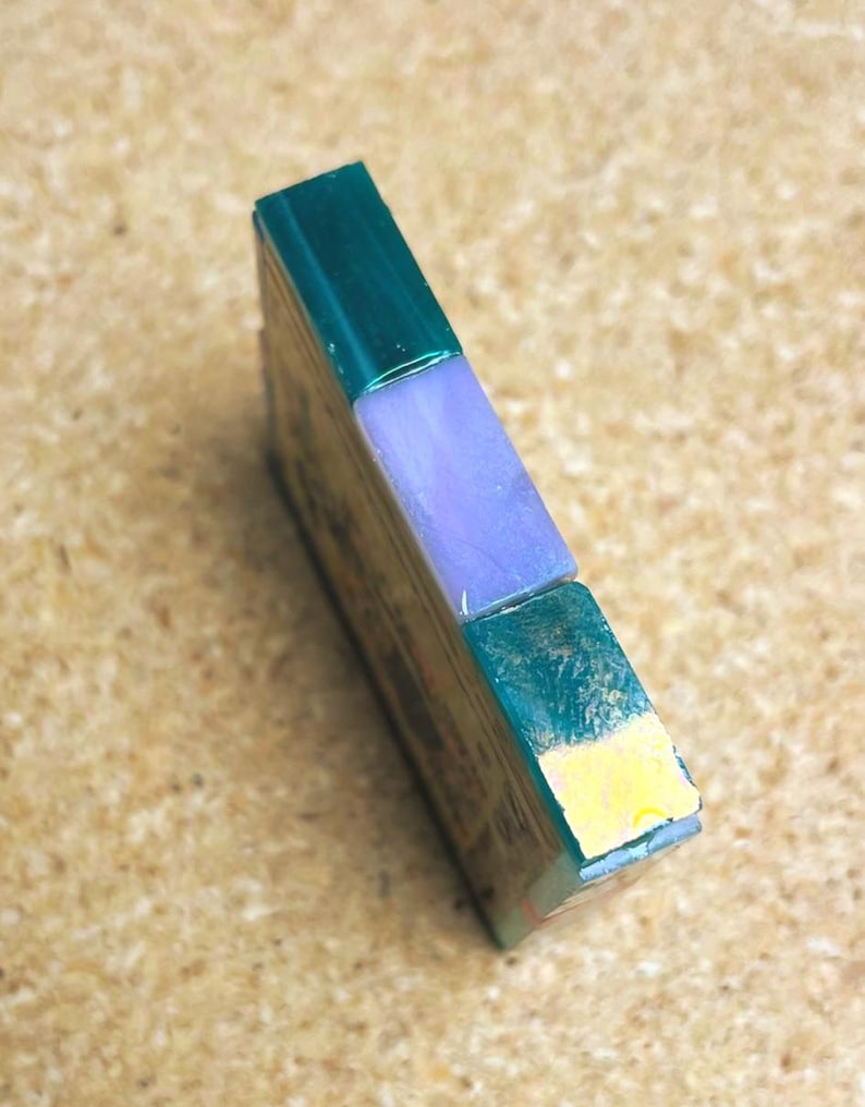 May include: A rectangular block featuring a dark green top section, a light purple middle segment, and a dark green bottom section with a small yellow accent on the lower edge. The object has a smooth, polished finish and a flat, vertical orientation.