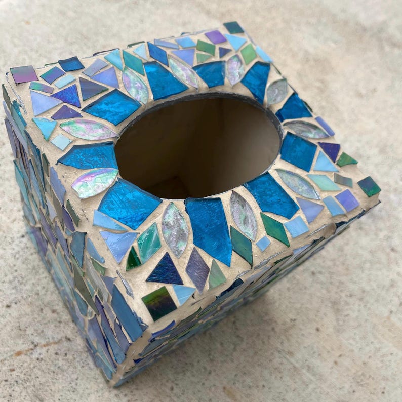 Op de afbeelding: Een vierkante tissueboxhouder versierd met een moza&iuml;ek van glasstukjes in diverse tinten blauw, groenblauw en iriserend wit. De glasfragmenten zijn ingebed in een lichtgekleurde voegmortel en hebben een ovale opening aan de bovenzijde.