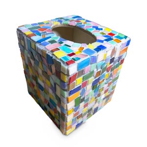 May include: A square tissue box cover decorated with a multi-colored mosaic pattern. The surface features small, irregularly shaped glass tiles in shades of blue, yellow, pink, green, orange, and red, set against a light-colored grout base.