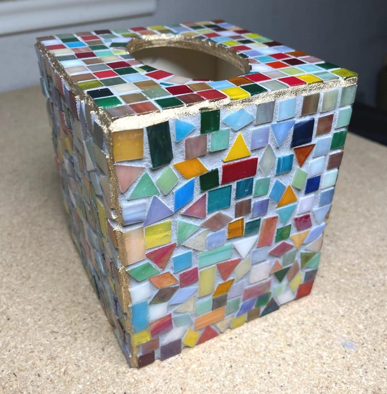 K&ouml;nnte beinhalten: Ein quadratischer Taschentuchbox-&Uuml;berzug, verziert mit einem bunten Mosaik aus kleinen Glasfliesen in verschiedenen geometrischen Formen. Die Kanten sind mit einer goldfarbenen Umrandung versehen und die Oberseite hat eine runde &Ouml;ffnung.