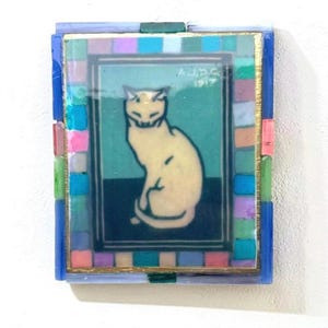 May include: A rectangular wall art piece featuring a central illustration of a cream-colored cat sitting on a dark surface against a teal background. The text A.J.D.G 1917 is visible. The frame is decorated with a mosaic of small, multi-colored glass tiles.