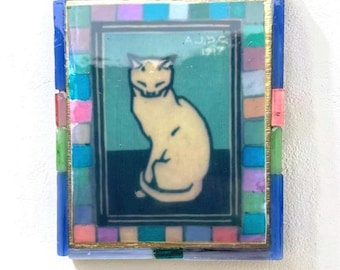 May include: A rectangular wall art piece featuring a central illustration of a cream-colored cat sitting on a dark surface against a teal background. The text A.J.D.G 1917 is visible. The frame is decorated with a mosaic of small, multi-colored glass tiles.