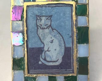 May include: A rectangular mosaic frame featuring a central illustration of a seated cat in shades of blue and purple. The border is composed of small, square tiles in iridescent white, dark green, and light blue, all outlined in gold.