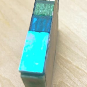 Pode incluir: Um bloco retangular com uma pilha vertical de segmentos coloridos em tons de verde brilhante, verde floresta, azul profundo, ciano, verde menta claro e azul marinho escuro. O objeto tem um acabamento liso e reflexivo e estrutura s&oacute;lida.