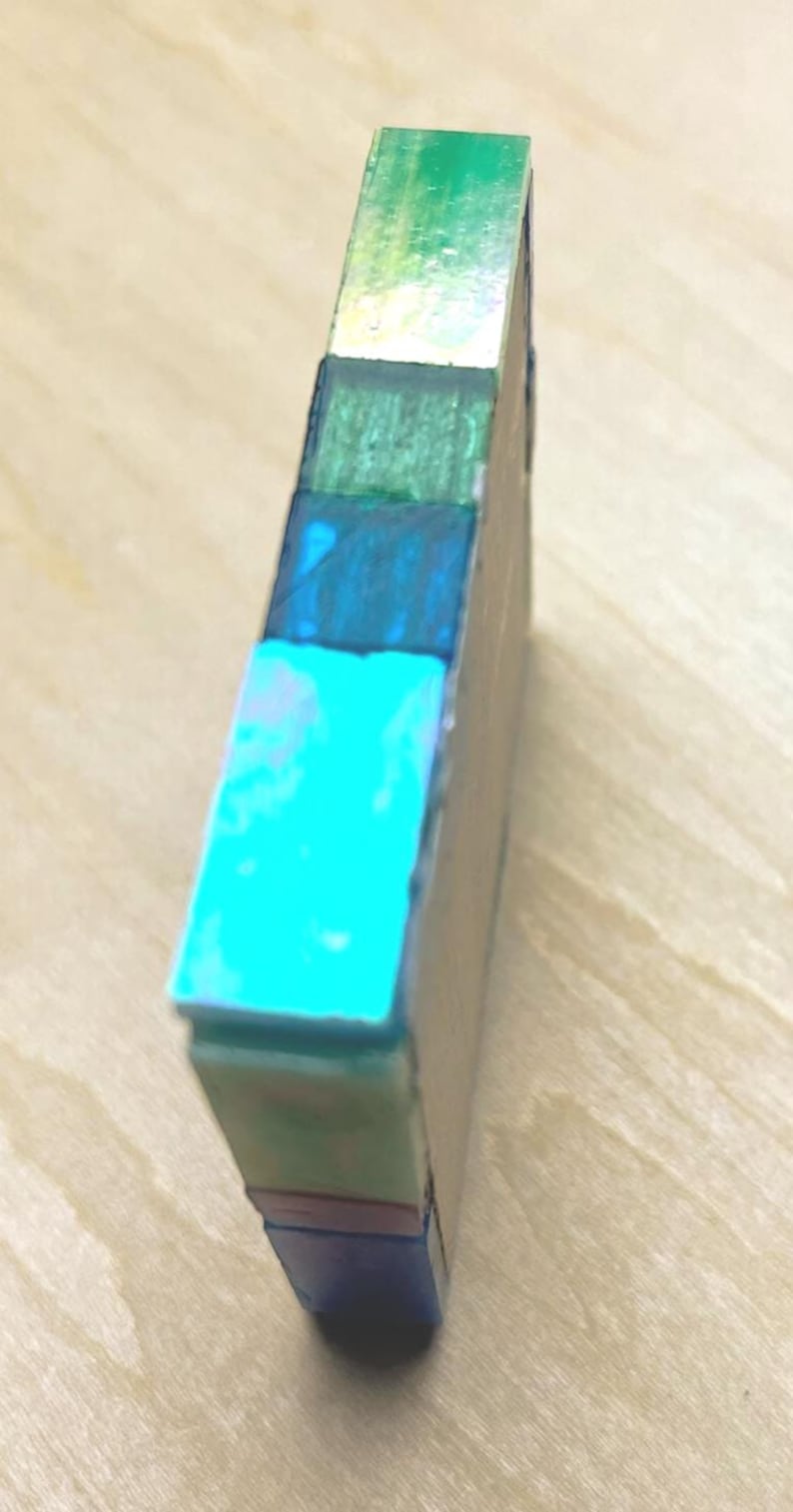 Pode incluir: Um bloco retangular com uma pilha vertical de segmentos coloridos em tons de verde brilhante, verde floresta, azul profundo, ciano, verde menta claro e azul marinho escuro. O objeto tem um acabamento liso e reflexivo e estrutura s&oacute;lida.