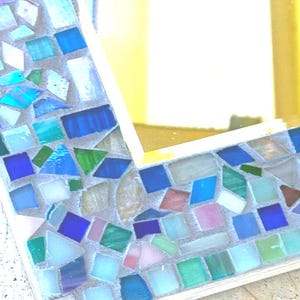 Op de afbeelding: Een rechthoekige spiegel met een decoratieve lijst bestaande uit diverse kleine, onregelmatig gevormde glastegels in tinten blauw, groen, wit en roze, geplaatst in lichtgekleurd voegmiddel rondom een reflecterend glazen middenpaneel.