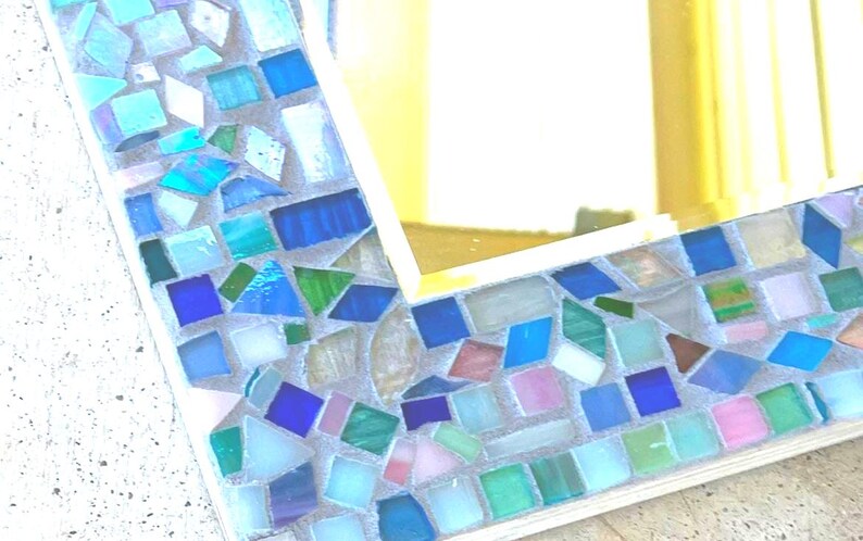 Op de afbeelding: Een rechthoekige spiegel met een decoratieve lijst bestaande uit diverse kleine, onregelmatig gevormde glastegels in tinten blauw, groen, wit en roze, geplaatst in lichtgekleurd voegmiddel rondom een reflecterend glazen middenpaneel.