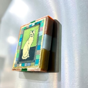 May include: A rectangular refrigerator magnet featuring a central illustration of a seated cat on a green and black background. The image is framed by a border of small, multicolored square tiles in shades of blue, green, and gray, with gold accents.