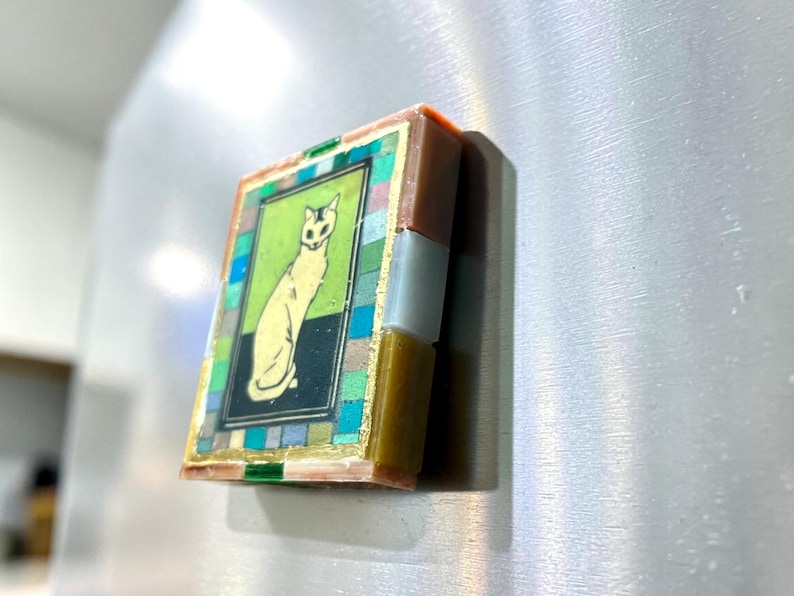 May include: A rectangular refrigerator magnet featuring a central illustration of a seated cat on a green and black background. The image is framed by a border of small, multicolored square tiles in shades of blue, green, and gray, with gold accents.