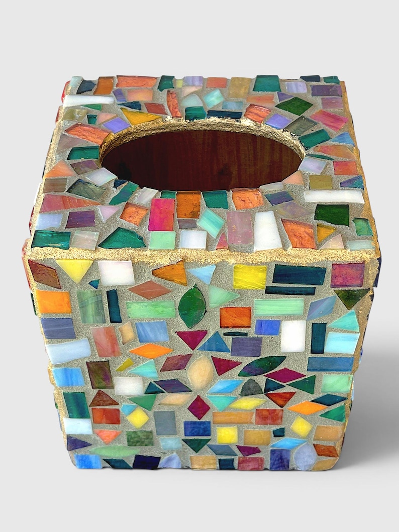 May include: A square tissue box cover featuring a multi-coloured mosaic design made of small, irregularly shaped glass pieces in shades of red, orange, yellow, green, blue, and purple, set in grey grout with a gold-painted top edge and opening.