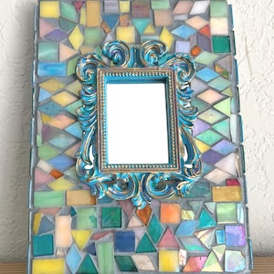 May include: A rectangular mirror frame decorated with a mosaic of multicolored glass tiles in various geometric shapes. Centered on the mosaic is a smaller, ornate, bright blue and gold-toned frame with intricate scrollwork detailing.