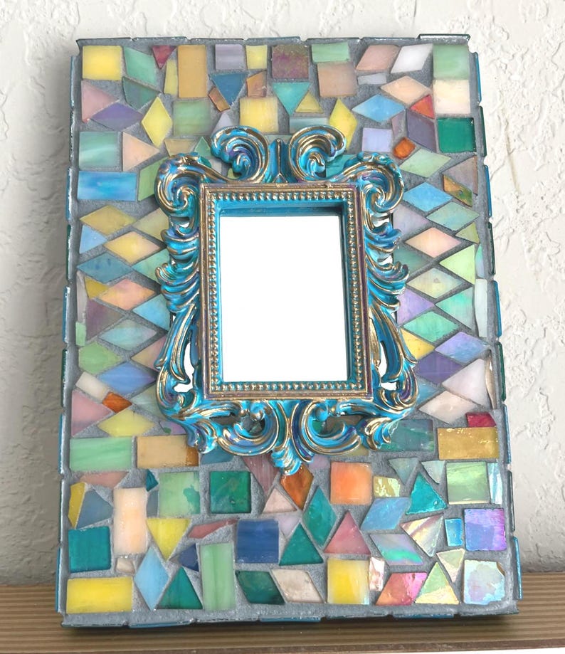 May include: A rectangular mirror frame decorated with a mosaic of multicolored glass tiles in various geometric shapes. Centered on the mosaic is a smaller, ornate, bright blue and gold-toned frame with intricate scrollwork detailing.