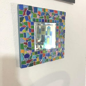May include: A square wall mirror featuring a mosaic frame composed of multi-colored glass shards in shades of blue, green, red, yellow, and pink, set in gray grout. The central reflective glass surface is square and surrounded by the mosaic border.