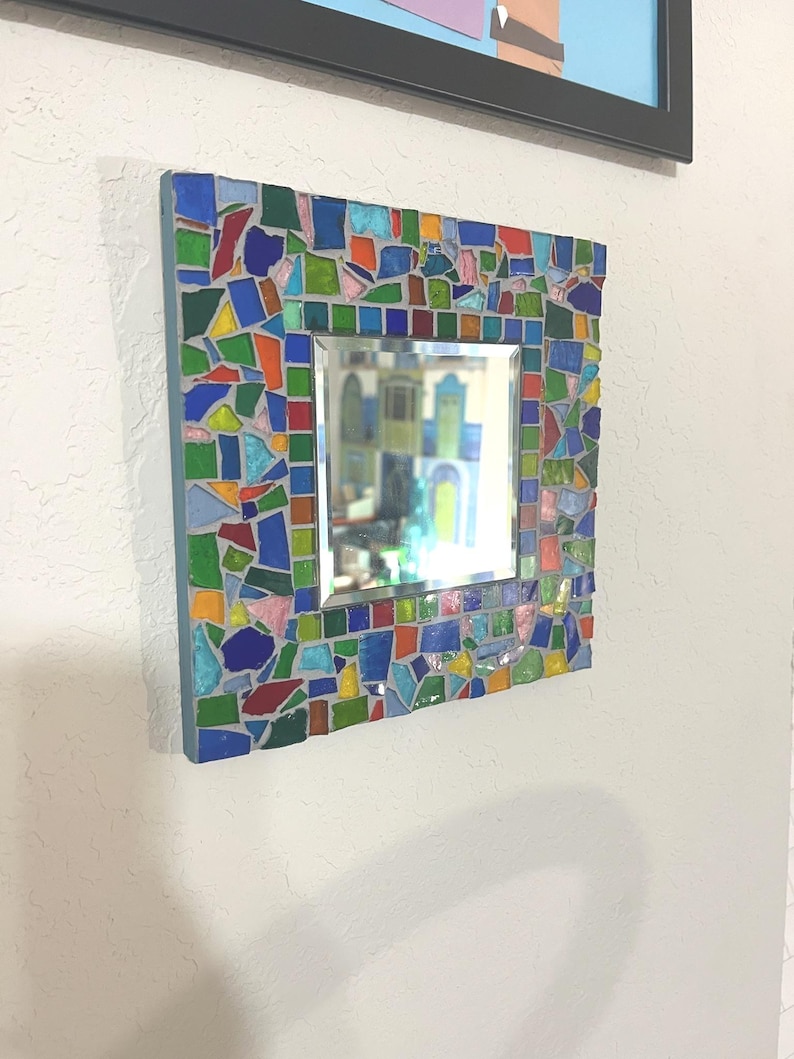 May include: A square wall mirror featuring a mosaic frame composed of multi-colored glass shards in shades of blue, green, red, yellow, and pink, set in gray grout. The central reflective glass surface is square and surrounded by the mosaic border.