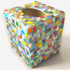 May include: A square tissue box cover featuring a multi-colored mosaic design. The exterior is covered in small, irregularly shaped glass tiles in shades of red, blue, green, yellow, orange, and purple, all set within a light gray grout.