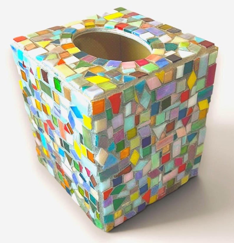 May include: A square tissue box cover featuring a multi-colored mosaic design. The exterior is covered in small, irregularly shaped glass tiles in shades of red, blue, green, yellow, orange, and purple, all set within a light gray grout.