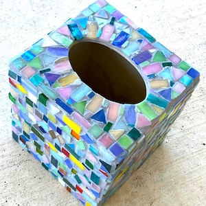 May include: A square tissue box cover decorated with a mosaic of small, multi-colored glass tiles. The tiles feature a variety of hues including pink, blue, green, yellow, and white, set into a light gray grout base with an oval opening.