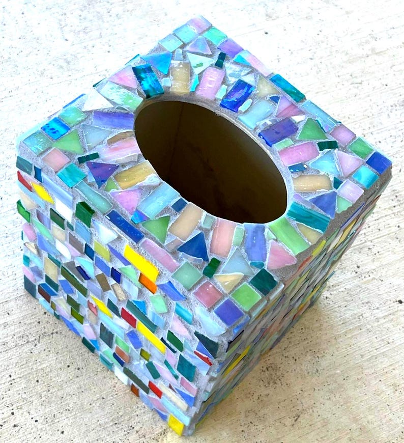 May include: A square tissue box cover decorated with a mosaic of small, multi-colored glass tiles. The tiles feature a variety of hues including pink, blue, green, yellow, and white, set into a light gray grout base with an oval opening.