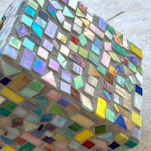 May include: A cube-shaped object covered in a mosaic of small, multi-colored glass tiles. The tiles feature various shapes including squares, rectangles, and triangles in shades of yellow, blue, green, pink, and iridescent finishes set in gray grout.