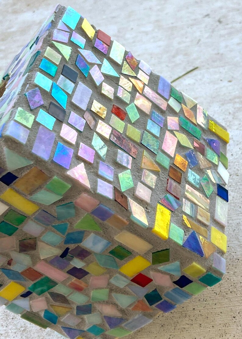 May include: A cube-shaped object covered in a mosaic of small, multi-colored glass tiles. The tiles feature various shapes including squares, rectangles, and triangles in shades of yellow, blue, green, pink, and iridescent finishes set in gray grout.