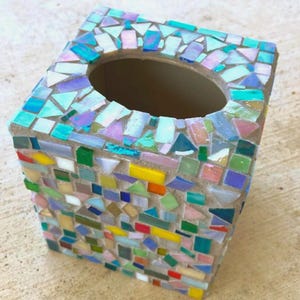 May include: A square tissue box cover decorated with a mosaic of small, multi-colored glass tiles in shades of blue, green, yellow, pink, and iridescent white. The tiles are arranged in a random pattern with visible grout between each piece.