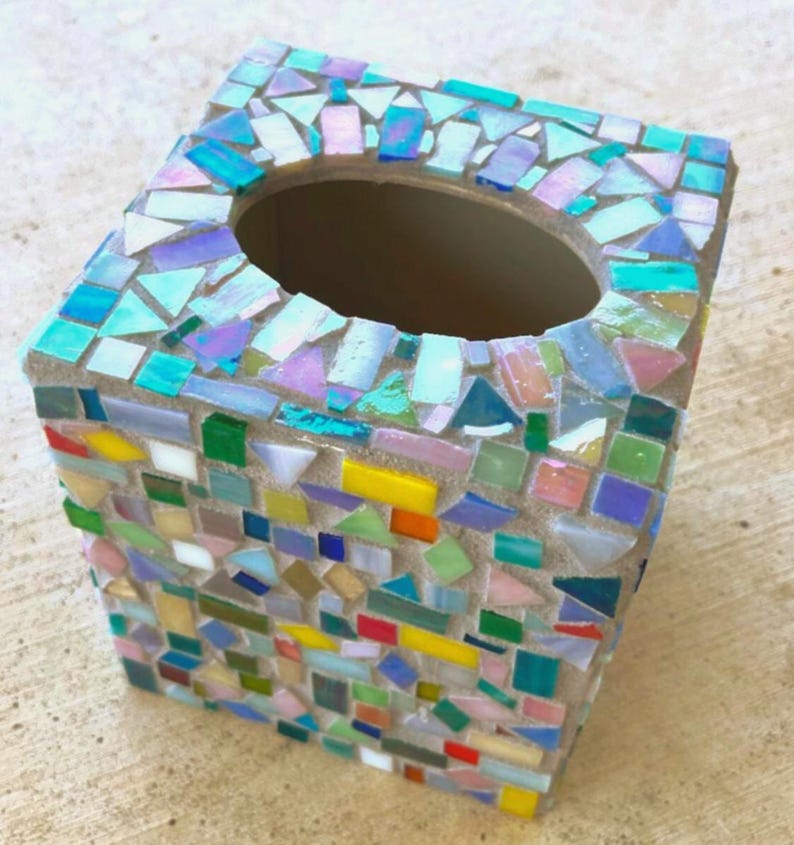 May include: A square tissue box cover decorated with a mosaic of small, multi-colored glass tiles in shades of blue, green, yellow, pink, and iridescent white. The tiles are arranged in a random pattern with visible grout between each piece.