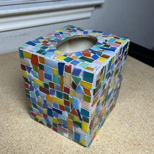May include: A square tissue box cover decorated with a multi-colored mosaic pattern of small, irregular glass tiles in shades of blue, green, yellow, orange, red, and white, featuring a circular opening on the top surface for dispensing tissues.