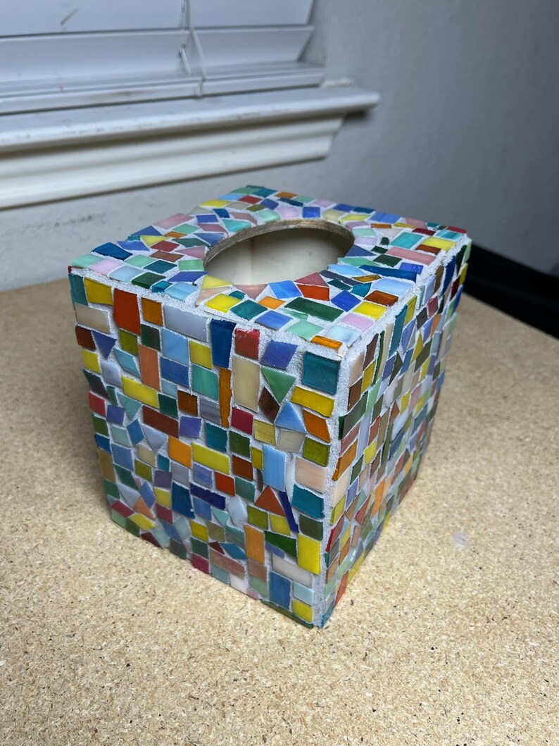 May include: A square tissue box cover decorated with a multi-colored mosaic pattern of small, irregular glass tiles in shades of blue, green, yellow, orange, red, and white, featuring a circular opening on the top surface for dispensing tissues.