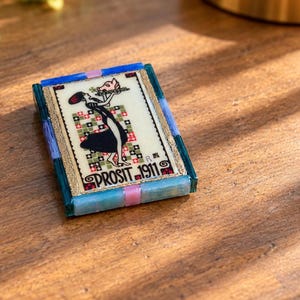 May include: A rectangular decorative tile featuring a central illustration of a figure holding a pig on a platter, set against a checkered background. The text PROSIT 1911 is printed below. The tile is framed by blue, teal, and pink glass segments.