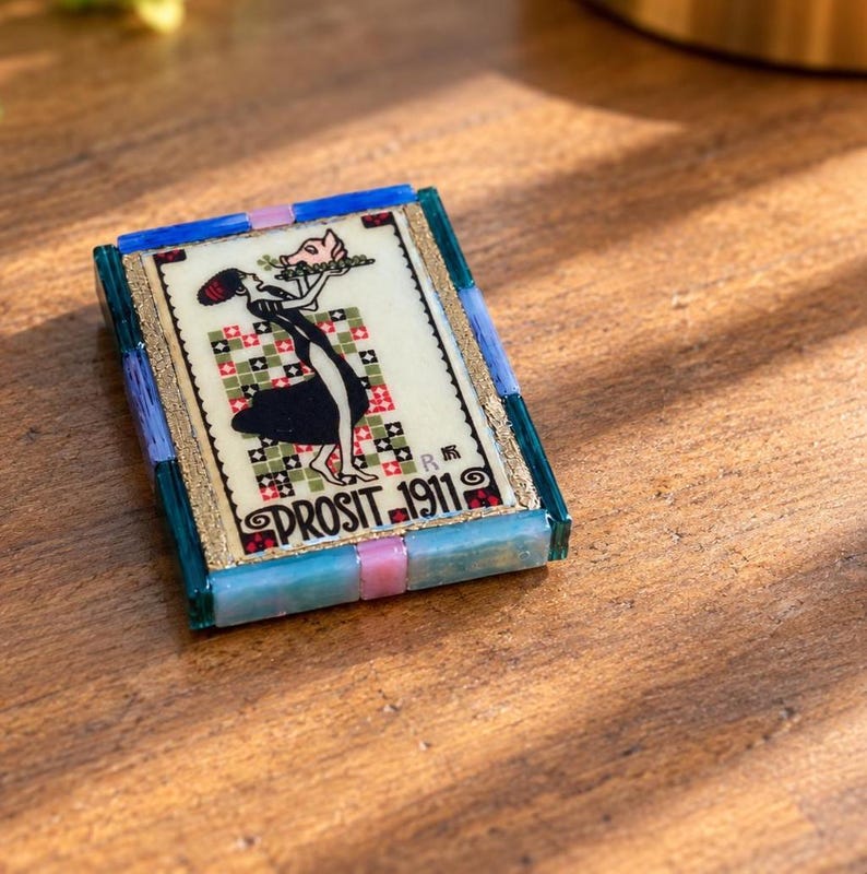 May include: A rectangular decorative tile featuring a central illustration of a figure holding a pig on a platter, set against a checkered background. The text PROSIT 1911 is printed below. The tile is framed by blue, teal, and pink glass segments.