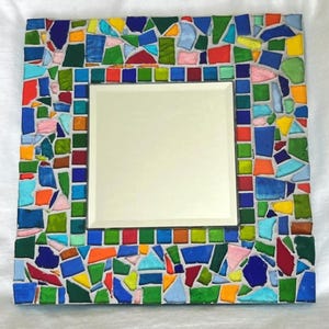 May include: A square mirror featuring a decorative frame composed of multi-colored glass mosaic tiles. The frame displays an array of irregular and square-shaped pieces in shades of blue, green, yellow, orange, red, and pink, set in white grout.
