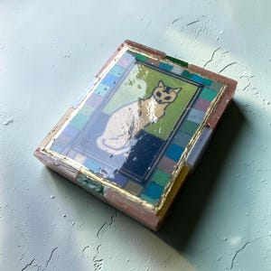 May include: A rectangular decorative object featuring a central illustration of a white cat on a green and dark blue background. The image is framed by a border of small, multicolored square tiles and finished with a glossy, reflective resin coating.