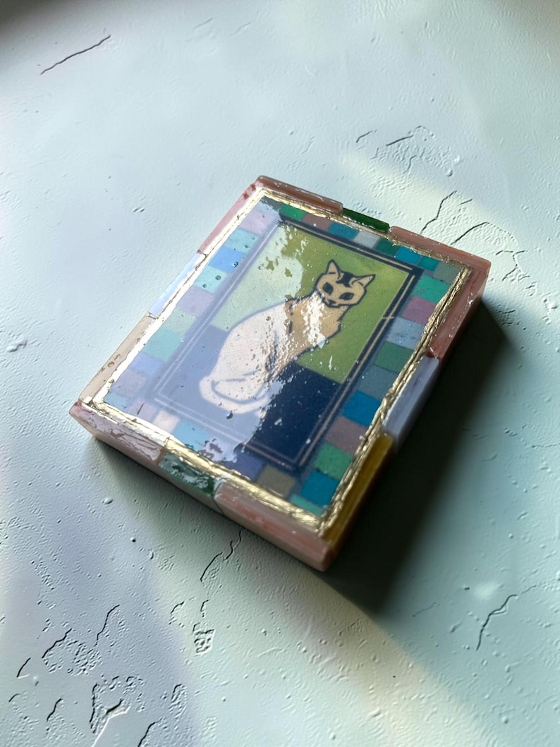 May include: A rectangular decorative object featuring a central illustration of a white cat on a green and dark blue background. The image is framed by a border of small, multicolored square tiles and finished with a glossy, reflective resin coating.