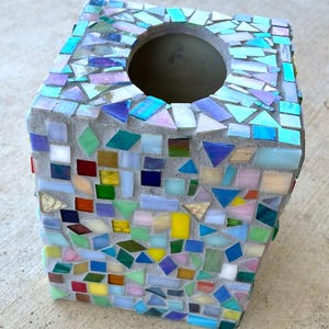 May include: A square tissue box cover featuring a mosaic design made of various small, multi-colored glass tiles embedded in gray grout. The tiles include shades of blue, green, yellow, red, and iridescent white, arranged in a random pattern.