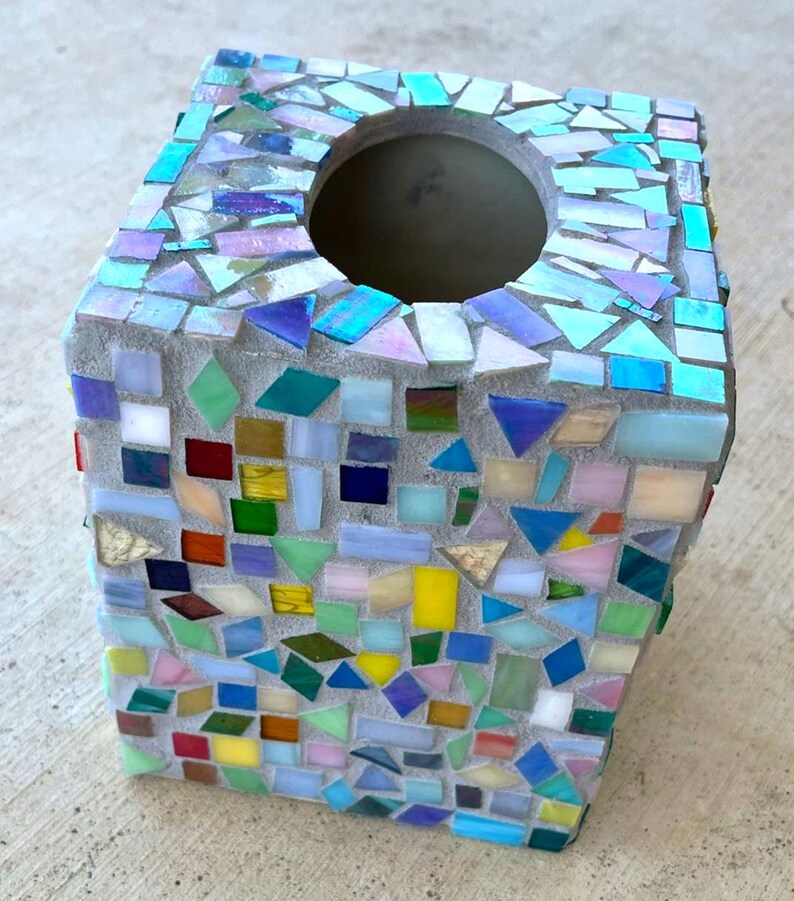 May include: A square tissue box cover featuring a mosaic design made of various small, multi-colored glass tiles embedded in gray grout. The tiles include shades of blue, green, yellow, red, and iridescent white, arranged in a random pattern.