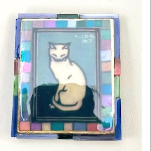 May include: A rectangular decorative tile featuring a central illustration of a seated white cat on a dark surface against a light blue background. The text A.J.D.G. 1917 is visible. The tile is bordered by a mosaic frame of small, multicolored glass squares.