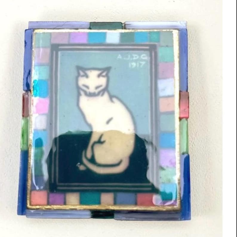 May include: A rectangular decorative tile featuring a central illustration of a seated white cat on a dark surface against a light blue background. The text A.J.D.G. 1917 is visible. The tile is bordered by a mosaic frame of small, multicolored glass squares.