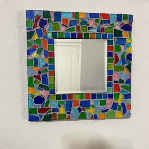 Op de afbeelding: Een vierkante wandspiegel met een brede lijst versierd met een kleurrijk mozaïek van onregelmatige glasscherven in blauw, groen, rood, geel en roze, gezet in grijze voegmortel, rondom een centraal vierkant reflecterend glazen oppervlak.