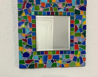 Puede incluir: Un espejo de pared cuadrado con un marco ancho decorado con un mosaico colorido de fragmentos de vidrio irregulares en tonos azul, verde, rojo, amarillo y rosa, fijados con lechada gris, rodeando una superficie de vidrio reflectante.